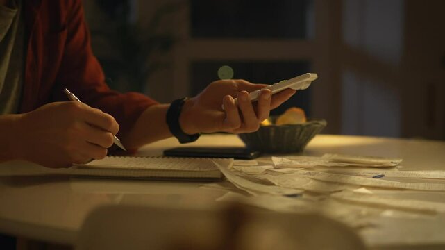 A person sits at a table in low light, jotting down notes with a pen while holding a calculator. Scattered financial receipts surround a bowl of fruit, creating a focused late-night atmosphere