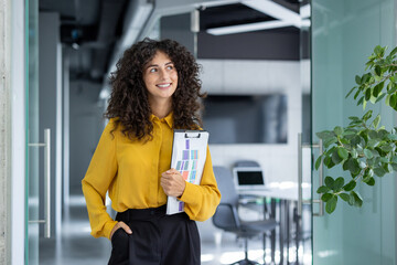 A confident businesswoman holding documents in a modern office setting. Her expression exudes...