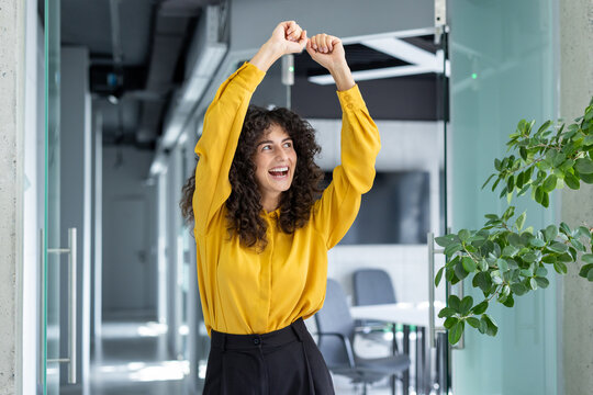 A joyful young woman celebrates with raised arms in a bright office. Her expression conveys excitement and accomplishment, epitomizing workplace success and positive energy.
