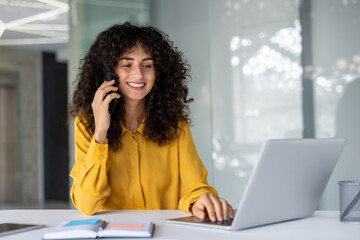 Smiling woman using phone and laptop at her desk, creating a productive and cheerful office...
