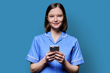 Young female nurse using smartphone, looking at camera on blue background