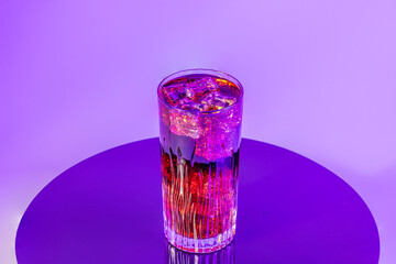 Transparent glass filled with sparkling red drink and ice cubes glowing under vibrant purple lighting on surface.