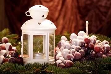 A white lantern standing on a green Christmas tree. Next to it are grapes covered in white icing. Christmas decorations in the warm light of candles. Horizontal framing.