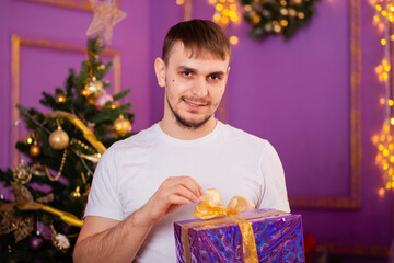 young man in white t-shirt and jeans on purple christmas background holding gift box . christmas tree with toys