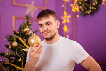 young man in white t-shirt and jeans on purple christmas background holding christmas tree toy in hands . christmas tree with toys