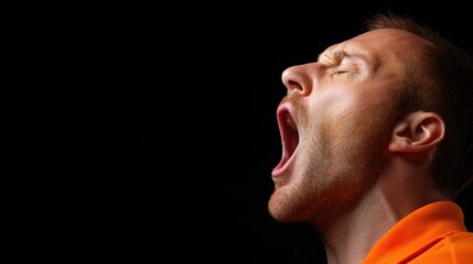 Obraz premium A man in an orange shirt expresses fatigue as he yawns widely, showcasing a moment of relaxation and tiredness with a dark backdrop emphasizing his features