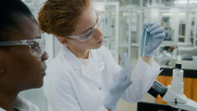 Close-up view of Black female chemist with digital tablet talking to Caucasian coworker who looking at content of test tube in laboratory