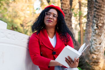 Teacher wearing glasses and a red beret relaxes on a white park bench, engrossed in a book and...