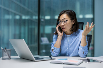 Asian businesswoman is engaged in a phone call while working at her laptop. She appears focused and is gesturing, suggesting a conversation about business matters.