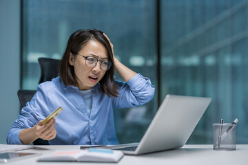 An Asian woman experiences frustration while using her laptop, holding a credit card in hand. The...