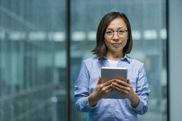 Asian woman confidently holds a tablet in a modern office. She appears professional, wearing...