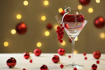 Creative presentation of Christmas cocktail in bauble and glass on white table against dark beige background with blurred lights. Space for text