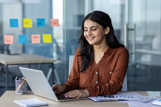 A cheerful woman, works on her laptop in a modern office. The setting features colorful sticky notes on glass and paperwork on the desk, highlighting productivity and professional focus.