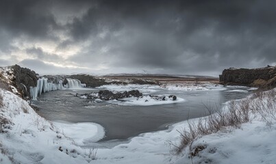 An overcast sky with snow on the ground and ice formations around the Goafoss waterfall in wintertime.