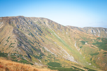 Naklejka premium Scenic view of mountain slopes bathed in soft sunlight, covered with green vegetation and rocky textures. A peaceful natural landscape showcasing the rugged beauty of high-altitude terrain