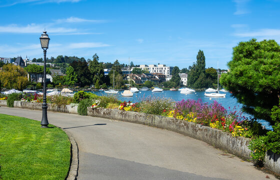Lakeside path in Morges lined with vibrant flowers, overlooking Geneva lake with boats and a  town in the background. 