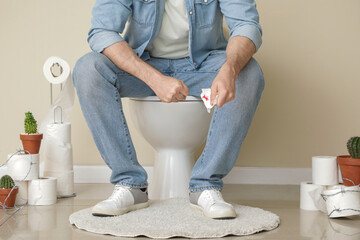 Suffering man with bloodstained paper sitting on toilet bowl in restroom. Hemorrhoid concept