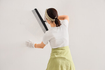 Young woman with putty knife working near light wall in room, back view