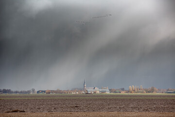 Dark storm clouds loom over a rural landscape with a church steeple in the distance during early morning hours
