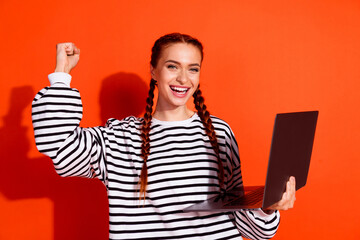 Young redhead woman in a striped pullover holding a laptop and expressing joy against a bright...