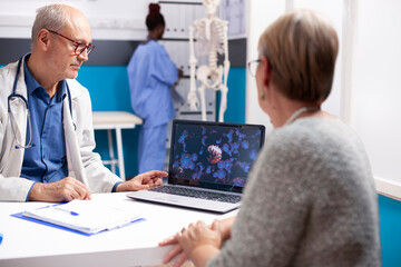 Obraz premium Senior male physician in lab coat discusses virus image on laptop with retired woman in well equipped hospital room. Old doctor and female patient looking at enlarged viral cells on digital device.