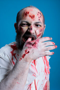 Portrait of insane psychotic zombie with blood on face biting his hand, feeling hungry for flesh. Close up shot of crazy starving creature chewing on own finger, isolated over studio background