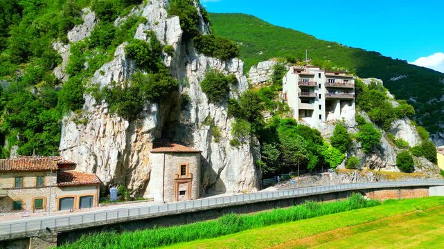 Impressive aerial truck right pan left push-in shot from Pioraco, the Eremo della Madonna della grotta in full display, nested inside the rugged rocky Sibillini Mountains with chunks of greenery, road