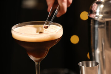 Bartender putting coffee bean into glass with delicious espresso martini against blurred lights, closeup