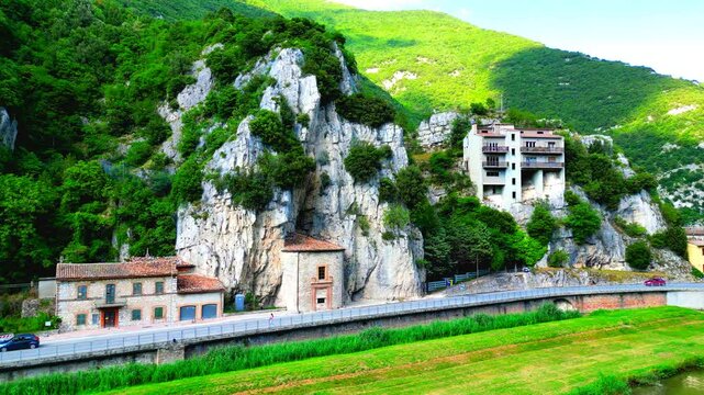 Inspiring aerial push-in shot of Pioraco, Potenza River, green meadows, road with cars, Eremo della Madonna della grotta, with the stunning display of the Sibillini mountains' rugged and rocky surface