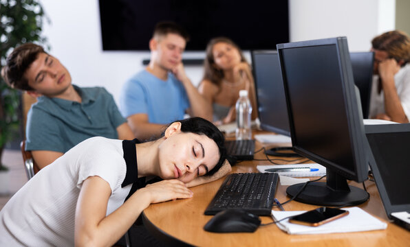 Young female developer tired after long workday napping near computer at office desk surrounded by coworkers..