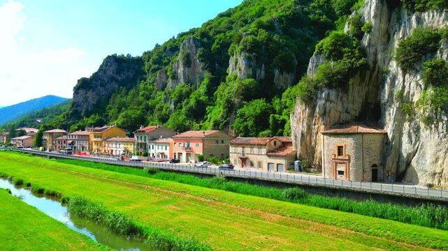 Calming aerial pedestal down shot from Pioraco, drone descending while capturing the mirroring waters of the Potenza River, the surrounding greenery, road with cars, Eremo della Madonna della grotta