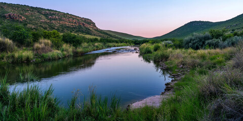 A peaceful river winding through a lush valley surrounded by steep green hills. The water reflects the soft hues of the evening sky.
