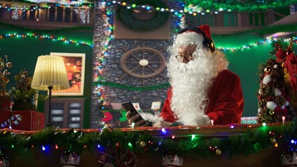 Cheerful Saint Nicholas listening to festive carols Christmas music on mobile phone, white bearded...