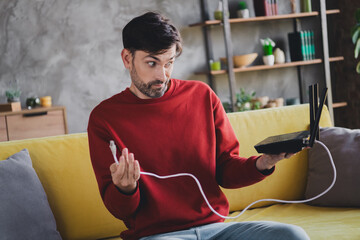 Young man puzzled with technology wearing casual red pullover indoors enjoying daylight in comfy...