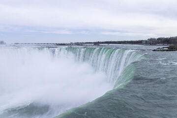 A close-up view of Niagara Falls (Horseshoe Falls) with powerful water flow, rising mist, and surrounding winter scenery under a cloudy sky.