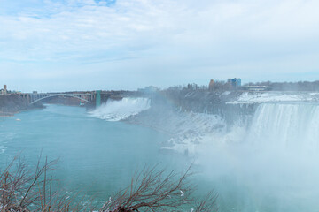 A winter view of Niagara Falls with mist rising, a faint rainbow, snow-covered cliffs, and the...