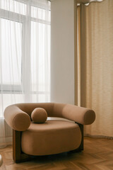Modern beige armchair with a minimalistic design, placed near a large window with sheer curtains, creating a calm and serene atmosphere in a cozy interior setting