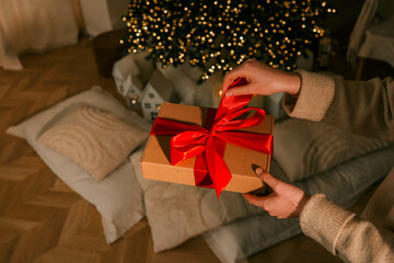 Person unwrapping a festive gift box with a red ribbon near a beautifully decorated Christmas tree....