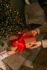 Hands exchanging a festive gift box with a red ribbon under a beautifully decorated Christmas tree....