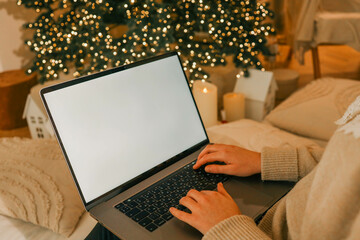 Close-up of a laptop with a blank white screen, hands typing on the keyboard, with a blurred background of a lit Christmas tree and festive decorations, creating a cozy holiday atmosphere