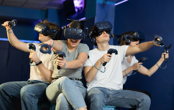 Cheerful emotional young girls and guys in VR headsets fully immersed in game, actively engaged in virtual reality world, manipulating objects or shooting targets with joysticks in hands