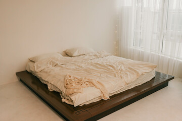 Minimalist bedroom with a cozy unmade bed, soft beige tones, and large windows draped with sheer...