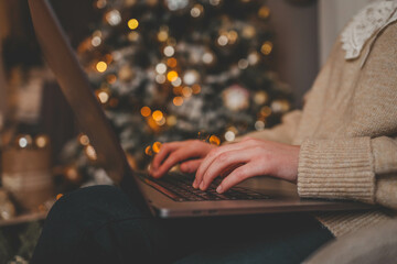 Hands typing on a laptop with a beautifully glowing Christmas tree in the background, creating a cozy and festive atmosphere indoors