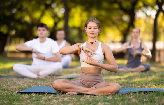 Young girl with closed eyes immersed in meditation during group yoga session in green sunlit summer park, sitting in lotus position, hands forming infinity sign with fingers folded in gyan mudra - Powered by Adobe