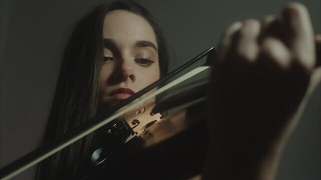 Closeup portrait of a beautiful young female violin player wearing a glitter black evening dress elegantly playing fiddle in a dark and hazy indoor environment