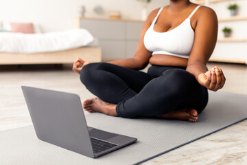Fototapeta premium Plus size black woman having online meditation or yoga class, sitting in lotus pose next to laptop at home, cropped view. Unrecognizable overweight young lady doing breathing exercises indoors