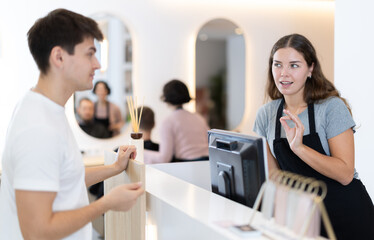 Female stylist stands near barbershop reception desk, communicates with male visitor, offer various hair care procedures, signs up client to visit stylist. Barbershop customer service in background