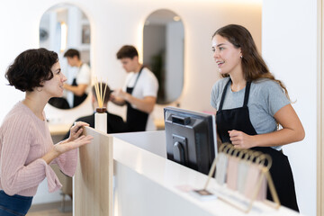 Female stylist stands near barbershop reception desk, communicates with woman visitor, offer...