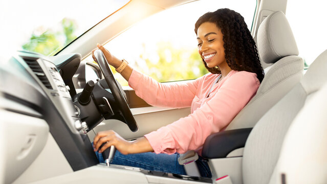 Riding In The City. Profile side view of happy black woman sitting in modern vehicle, driving her new auto, holding steering wheel and gear shift. Experienced female driver enjoying trip and travel