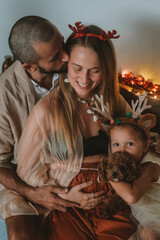 Obraz premium Family Christmas portrait. Pregnant woman with her husband and little daughter sitting on the couch and hugging. Their toy poodle next to them. Holiday lights in the background. Love and tenderness.